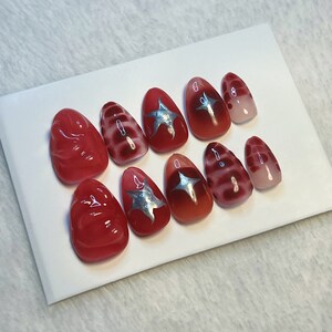 May include: Set of ten red and silver press-on nails with different designs. Some nails have a swirl pattern, some have a star, and some are solid red.