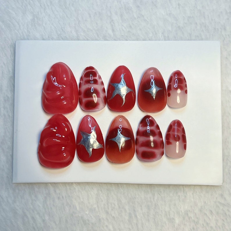 May include: Set of ten red and silver press-on nails with different designs. Some nails have a swirl design, some have a star design, and some have a striped design.