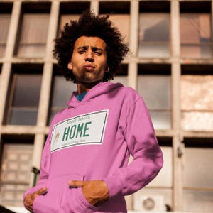 May include: A pink hoodie with a white graphic of a Saskatchewan licence plate that says "HOME Land of Living Skies".