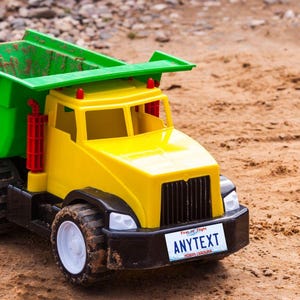 Pode incluir: Um caminhão de brinquedo amarelo com uma caçamba verde e rodas pretas. O caminhão tem uma placa que diz "ANYTEXT" com "First Class Flight" e "NORTH CAROLINA" impresso nela.