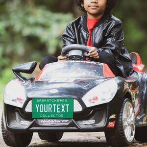 May include: A child wearing a black leather jacket drives a black and red toy car with a green number plate that reads "Saskatchewan YOURTEXT COLLECTOR".