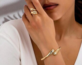 May include: Gold-toned jewelry set featuring a ring and bracelet. The ring wraps around the finger with two rounded shapes. The bracelet has a similar design, with two curved, teardrop-shaped ends that meet on the wrist. The jewelry has a polished finish.