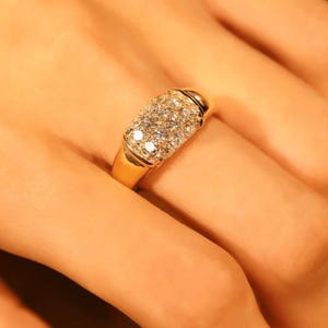 May include: A gold ring with a rectangular face set with numerous small, sparkling clear stones. The ring is on a finger, and the background is a warm, neutral tone. The ring is made of S925 sterling silver.