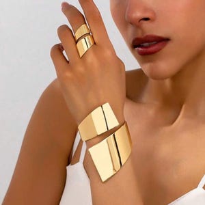 May include: Gold-toned jewelry set featuring a ring and a cuff bracelet. The ring has a split design, and the cuff bracelet wraps around the wrist. Both pieces have a geometric, angular shape and a polished finish.
