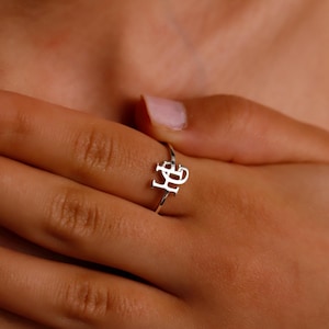 May include: A silver ring with the letters "HB" on it. The ring is worn on the ring finger of a person's hand.