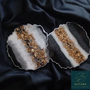 May include: Two circular resin coasters with irregular, wavy edges. The design features a black and white marbled pattern with a prominent central band of textured gold leaf flakes. A logo in the corner displays the text ALYAMA SWEETGLOWRESIN.