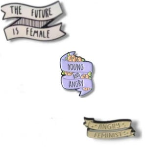 Può includere: Tre spille smaltate con slogan femministi. Una spilla recita "THE FUTURE IS FEMALE" su uno striscione bianco. Un'altra recita "YOUNG AND ANGRY" su uno striscione lavanda con accenti floreali. La terza spilla dice "ANGRY FEMINIST" su uno striscione color cachi.