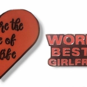 Op de afbeelding: Twee emaille pins. Eén is hartvormig, rood met zwarte tekst die luidt "you are the love of my life". De andere is rechthoekig, rood met zwarte tekst die luidt "WORLD'S BEST EX GIRLFRIEND."