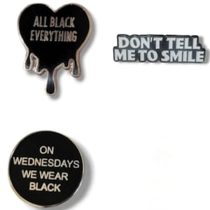 May include: Three black and silver enamel pins with white text. The first pin is a dripping heart shape with the text "ALL BLACK EVERYTHING". The second pin is rectangular with the text "DON'T TELL ME TO SMILE". The third pin is round with the text "ON WEDNESDAYS WE WEAR BLACK".