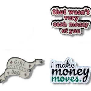 Può includere: Quattro spille smaltate con diversi detti. La prima spilla è bianca con testo nero che dice "that wasn't very cash money of you". La seconda spilla è bianca con testo nero che dice "girl boss" su uno stendardo bianco con accenti neri e rosa. La terza spilla è bianca con testo nero che dice "i make money moves" con accenti verdi. La quarta spilla è bianca con testo nero che dice "i make money moves" con accenti verdi.