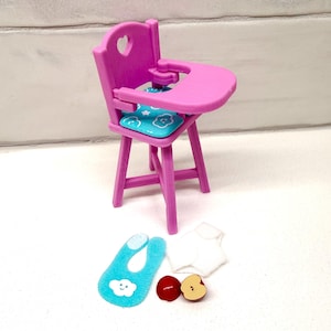 May include: A miniature pink high chair with a heart-shaped cutout in the backrest and a tray. It includes a blue seat cushion with cloud designs, a blue bib, a white shirt, and two apple slices.
