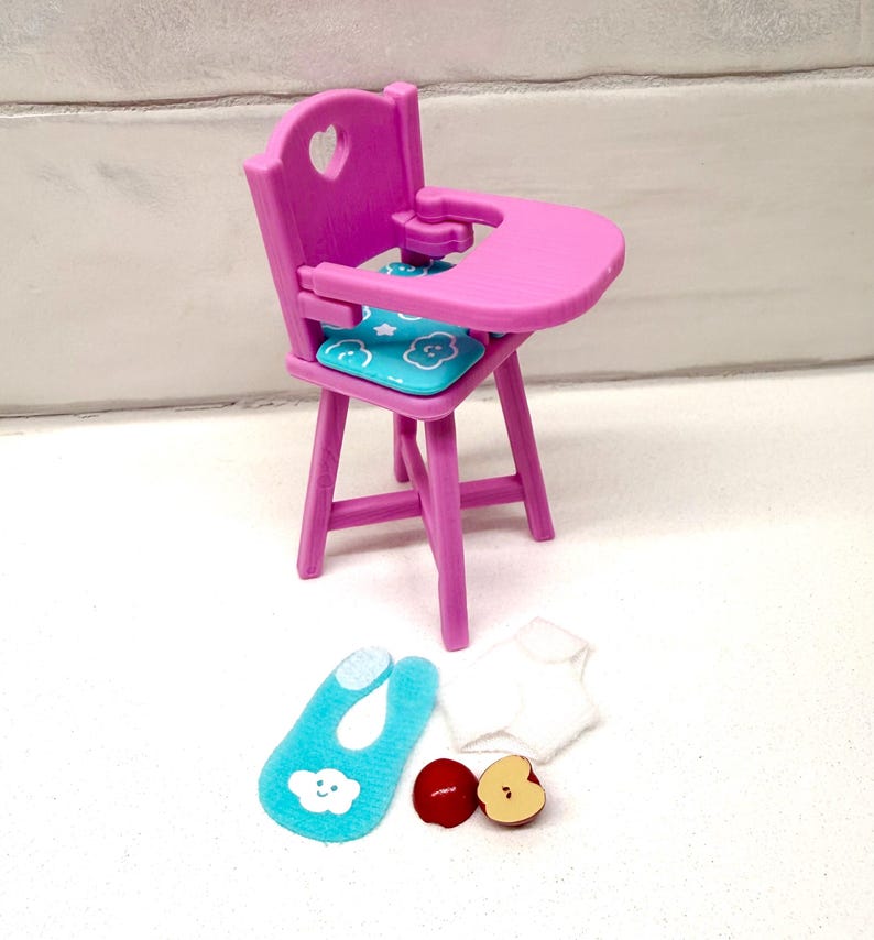 May include: A miniature pink high chair with a heart-shaped cutout in the backrest and a tray. It includes a blue seat cushion with cloud designs, a blue bib, a white shirt, and two apple slices.