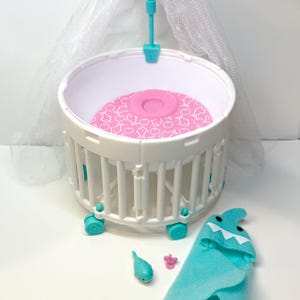 May include: A white and teal baby doll crib with a pink patterned mattress, a teal heart-topped canopy, and a teal shark-themed blanket. Also included are a teal dolphin and a pink pacifier.