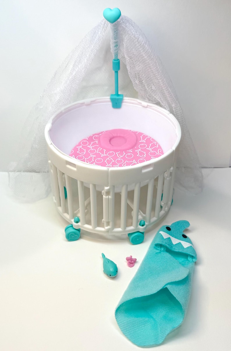 May include: A white and teal baby doll crib with a pink patterned mattress, a teal heart-topped canopy, and a teal shark-themed blanket. Also included are a teal dolphin and a pink pacifier.