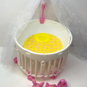 May include: A white plastic baby crib with a yellow mattress featuring a pink bunny pattern. The crib has a pink canopy with a heart-shaped topper. Includes pink bunny figures and a pink onesie.