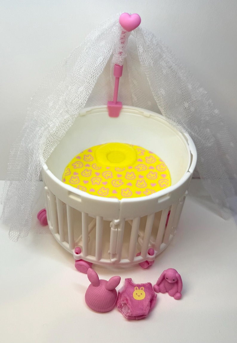 May include: A white plastic baby crib with a yellow mattress featuring a pink bunny pattern. The crib has a pink canopy with a heart-shaped topper. Includes pink bunny figures and a pink onesie.