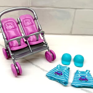 May include: A toy stroller with two pink seats, a silver frame, and pink wheels. Two blue baseball caps and two blue and white striped dresses with car graphics are also included.