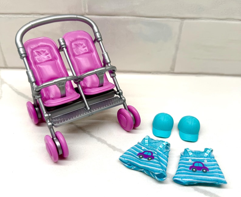 May include: A toy stroller with two pink seats, a silver frame, and pink wheels. Two blue baseball caps and two blue and white striped dresses with car graphics are also included.