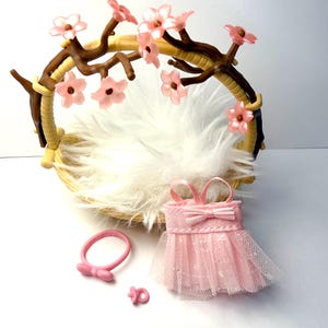 May include: A newborn photography prop set featuring a woven basket with a faux fur lining, adorned with a floral branch. Includes a pink dress, headband, and pacifier. The dress has a bow and tulle skirt.