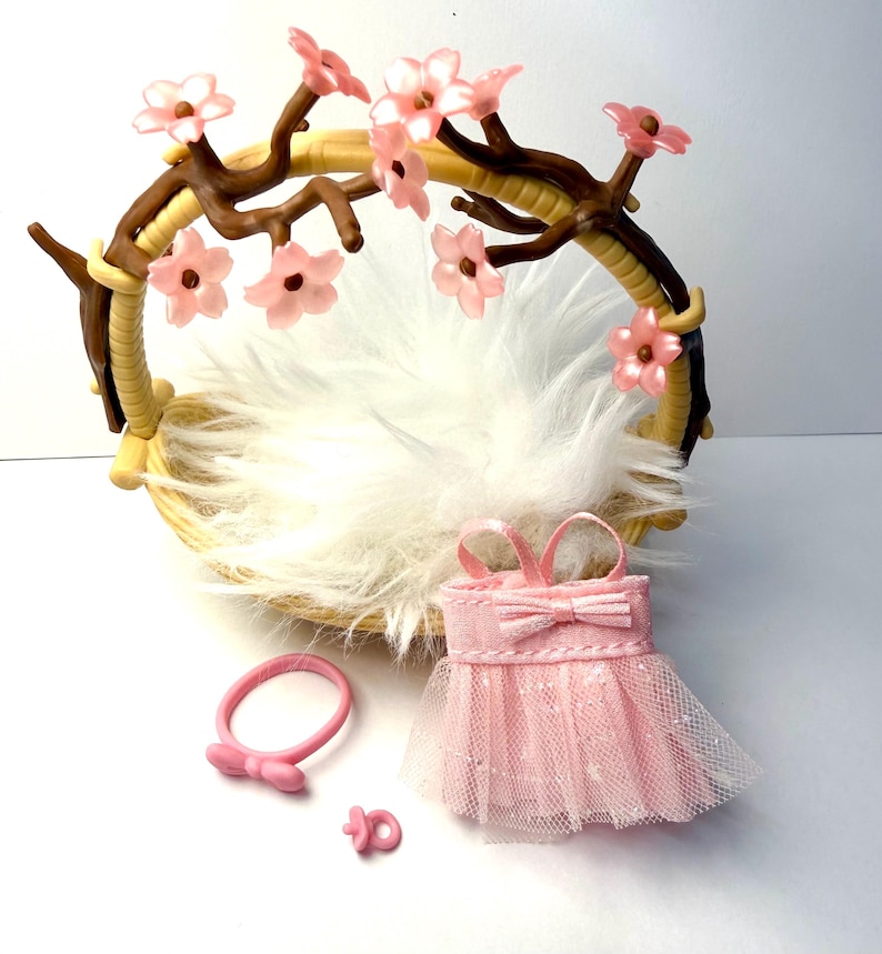 May include: A newborn photography prop set featuring a woven basket with a faux fur lining, adorned with a floral branch. Includes a pink dress, headband, and pacifier. The dress has a bow and tulle skirt.