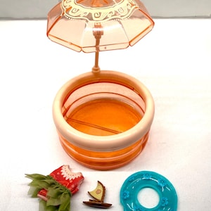 May include: A doll-sized playset featuring a peach-colored pool with a matching umbrella, a red and green dress, a small wooden boat, and a turquoise pool float with floral accents. The pool is round and has a light peach rim.