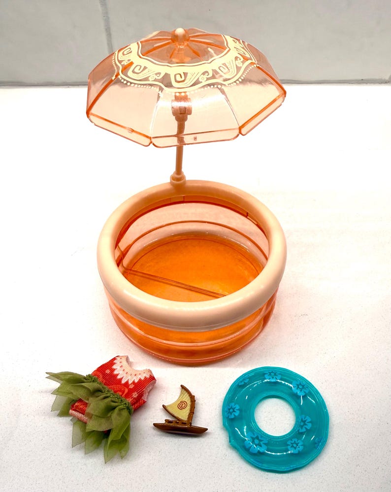 May include: A doll-sized playset featuring a peach-colored pool with a matching umbrella, a red and green dress, a small wooden boat, and a turquoise pool float with floral accents. The pool is round and has a light peach rim.