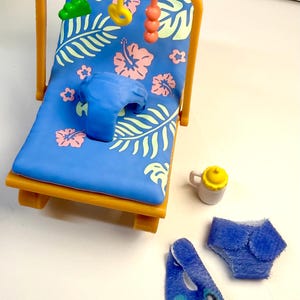 May include: A miniature baby beach chair with a blue floral print, a blue pillow, and hanging toys. Accessories include a baby bottle, diaper, and a blue bib with a cartoon character design.