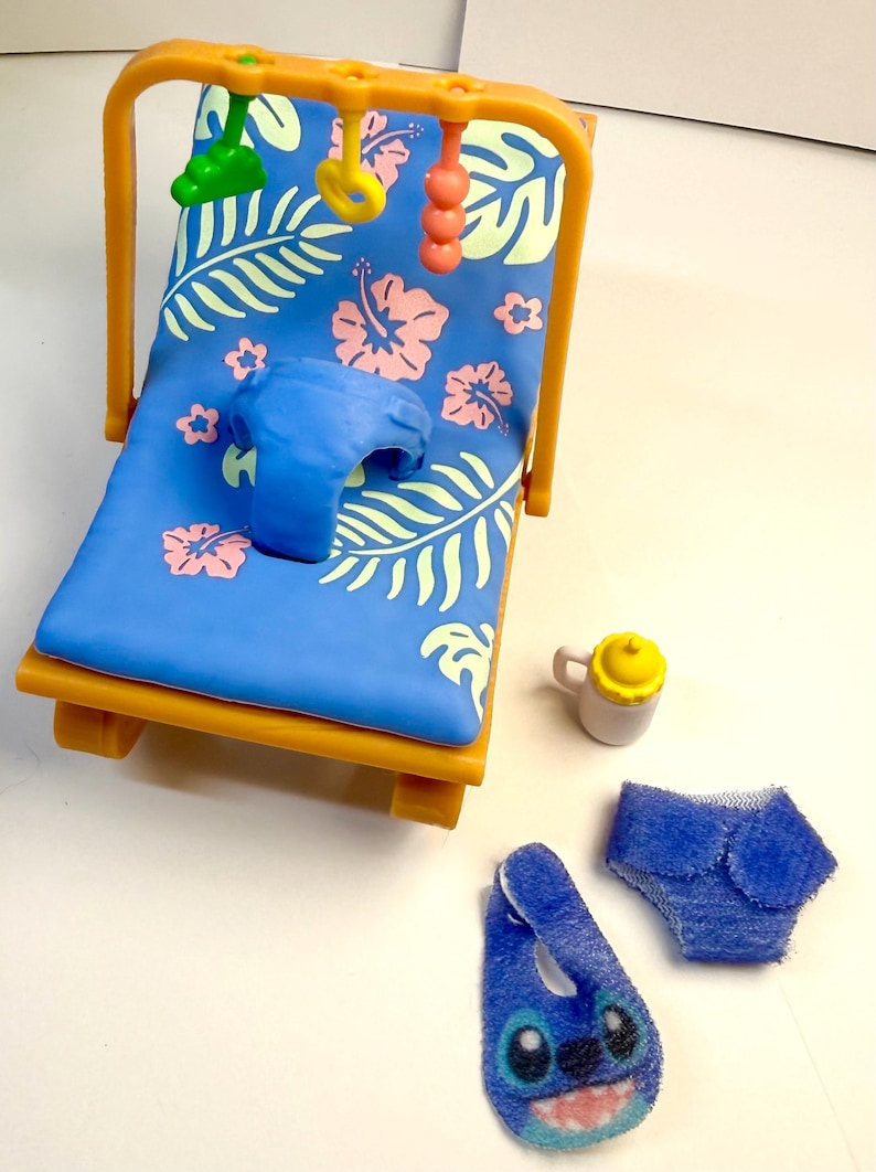 May include: A miniature baby beach chair with a blue floral print, a blue pillow, and hanging toys. Accessories include a baby bottle, diaper, and a blue bib with a cartoon character design.