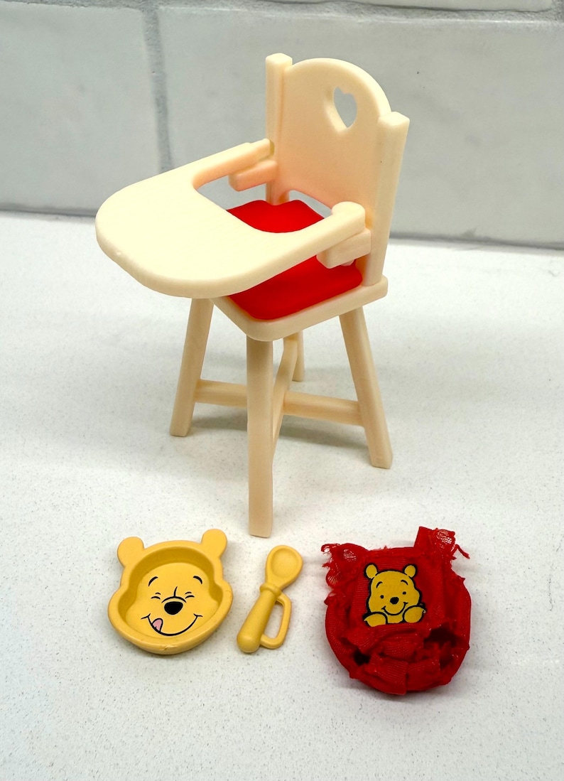 May include: A miniature Winnie the Pooh themed high chair set. The set includes a cream-colored high chair with a red seat, a yellow bowl, a yellow spoon, and a red bib. The bowl and bib feature Winnie the Pooh's face.