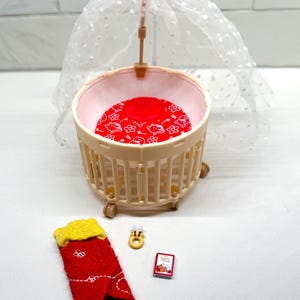 May include: A miniature baby crib with a red patterned mattress and a sheer canopy. Accessories include a red and yellow sock, a small book, and a yellow bee-shaped ring. The crib is beige and has small wheels.