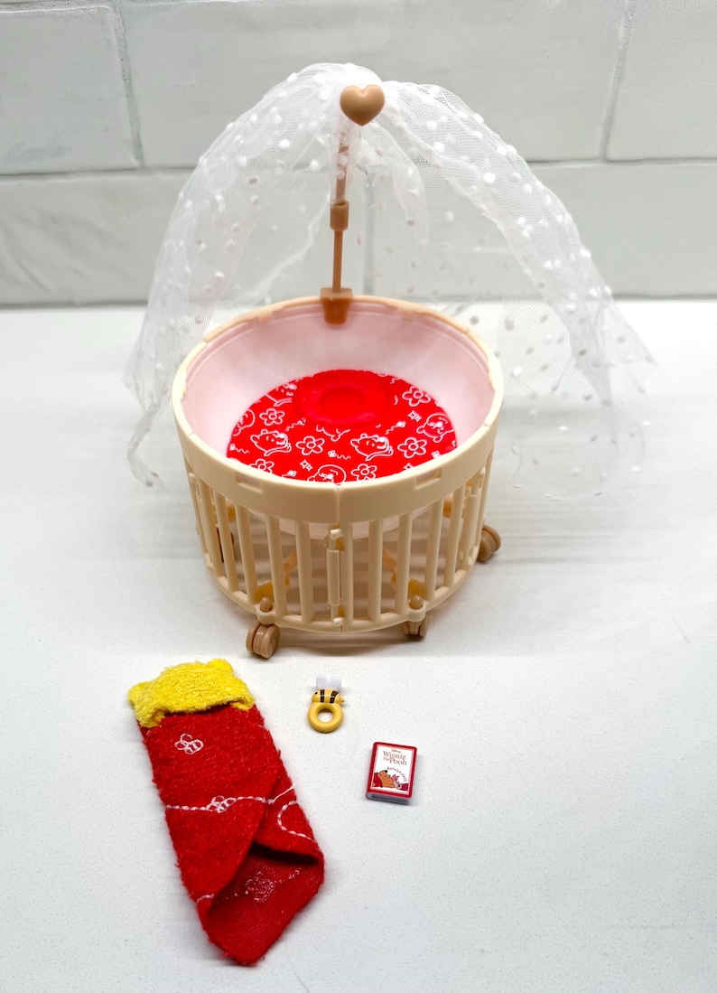 May include: A miniature baby crib with a red patterned mattress and a sheer canopy. Accessories include a red and yellow sock, a small book, and a yellow bee-shaped ring. The crib is beige and has small wheels.