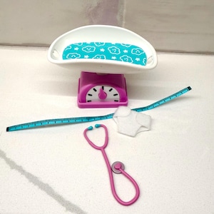 May include: A pink and white toy baby scale with a blue cloud-patterned tray, a blue measuring tape, a pink stethoscope, and a white diaper. The scale has a dial and a curved tray.