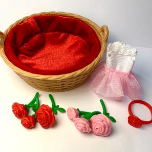 May include: A wicker basket with a red satin lining, a white and pink dress, red and pink roses, and a red ring with a rose. The items are likely doll accessories.