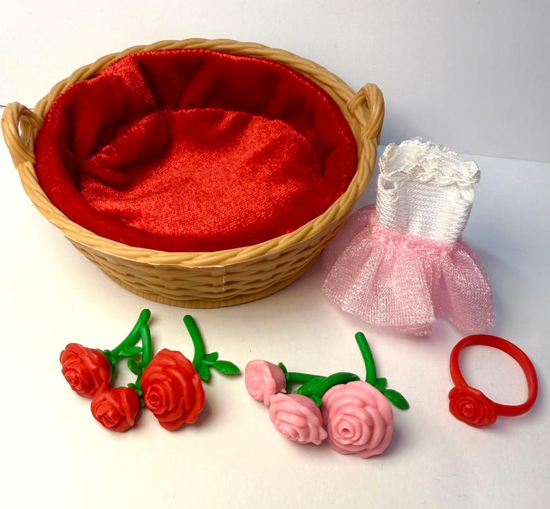 May include: A wicker basket with a red satin lining, a white and pink dress, red and pink roses, and a red ring with a rose. The items are likely doll accessories.