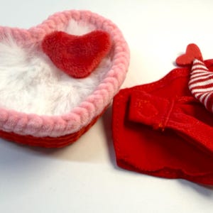May include: A heart-shaped, red and pink basket with a fluffy white interior and a small red heart. Next to it is a red fabric item with a small hat with red and white stripes.