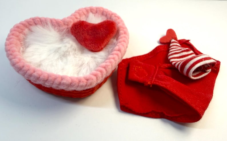 May include: A heart-shaped, red and pink basket with a fluffy white interior and a small red heart. Next to it is a red fabric item with a small hat with red and white stripes.