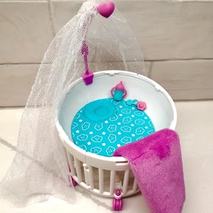 May include: A white plastic doll crib with a blue patterned mattress, a pink blanket, and a sheer canopy. The crib has a pink heart-shaped accent and pink wheels. A blue baby bottle and a pink flower are also inside the crib.