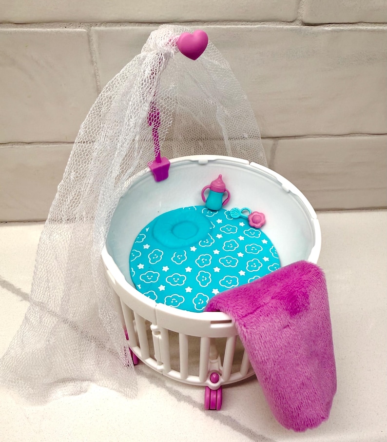 May include: A white plastic doll crib with a blue patterned mattress, a pink blanket, and a sheer canopy. The crib has a pink heart-shaped accent and pink wheels. A blue baby bottle and a pink flower are also inside the crib.