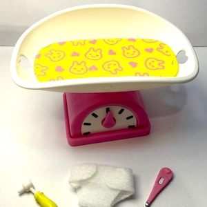 May include: A pink and white toy baby scale with a yellow patterned tray, a diaper, a thermometer, and a yellow otoscope. The tray has a yellow background with pink bunny and heart shapes.