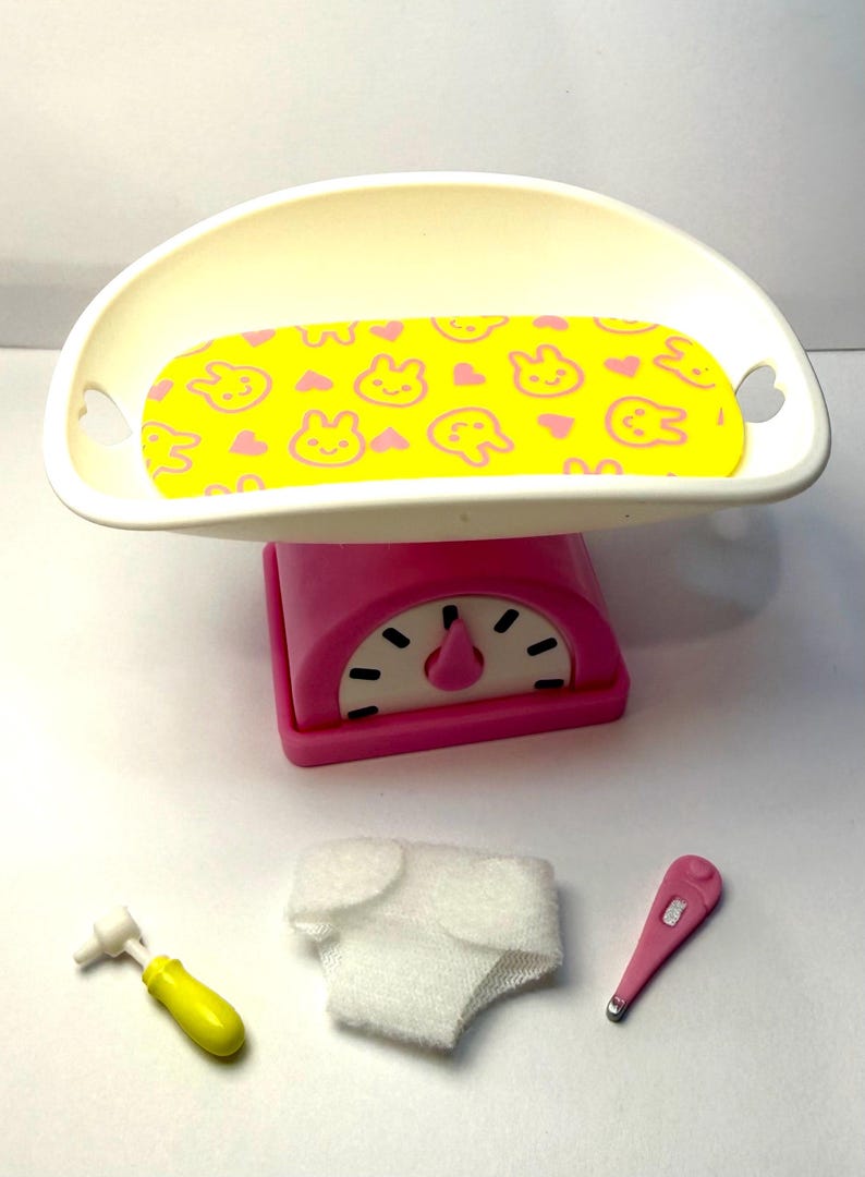 May include: A pink and white toy baby scale with a yellow patterned tray, a diaper, a thermometer, and a yellow otoscope. The tray has a yellow background with pink bunny and heart shapes.