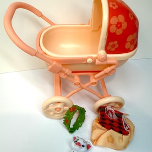 May include: A vintage toy baby carriage in shades of peach and cream, with a red floral-patterned canopy. Includes a small green wreath, a white pig figurine, and a blanket with a red and black plaid pattern.