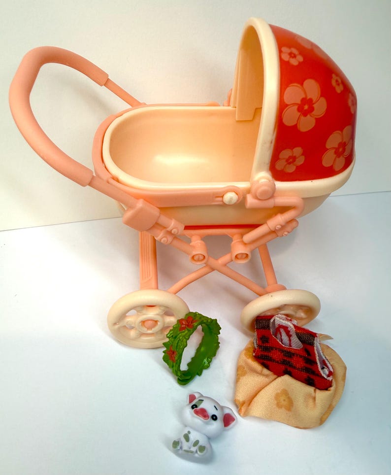 May include: A vintage toy baby carriage in shades of peach and cream, with a red floral-patterned canopy. Includes a small green wreath, a white pig figurine, and a blanket with a red and black plaid pattern.