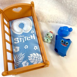 May include: A wooden doll crib with a blue Disney Stitch blanket and a white pillow. Includes a blue Stitch outfit, a blue headpiece, and a small green doll. The crib has a heart-shaped cutout.
