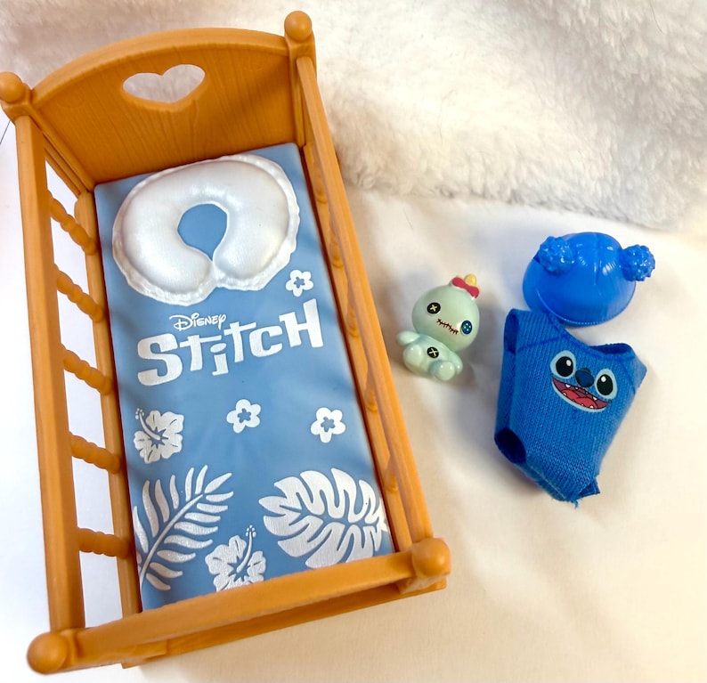 May include: A wooden doll crib with a blue Disney Stitch blanket and a white pillow. Includes a blue Stitch outfit, a blue headpiece, and a small green doll. The crib has a heart-shaped cutout.