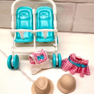 May include: A white and teal double doll stroller with matching teal wheels. Accessories include a pink gingham dress, a pink gingham sun hat, and two beige straw hats.