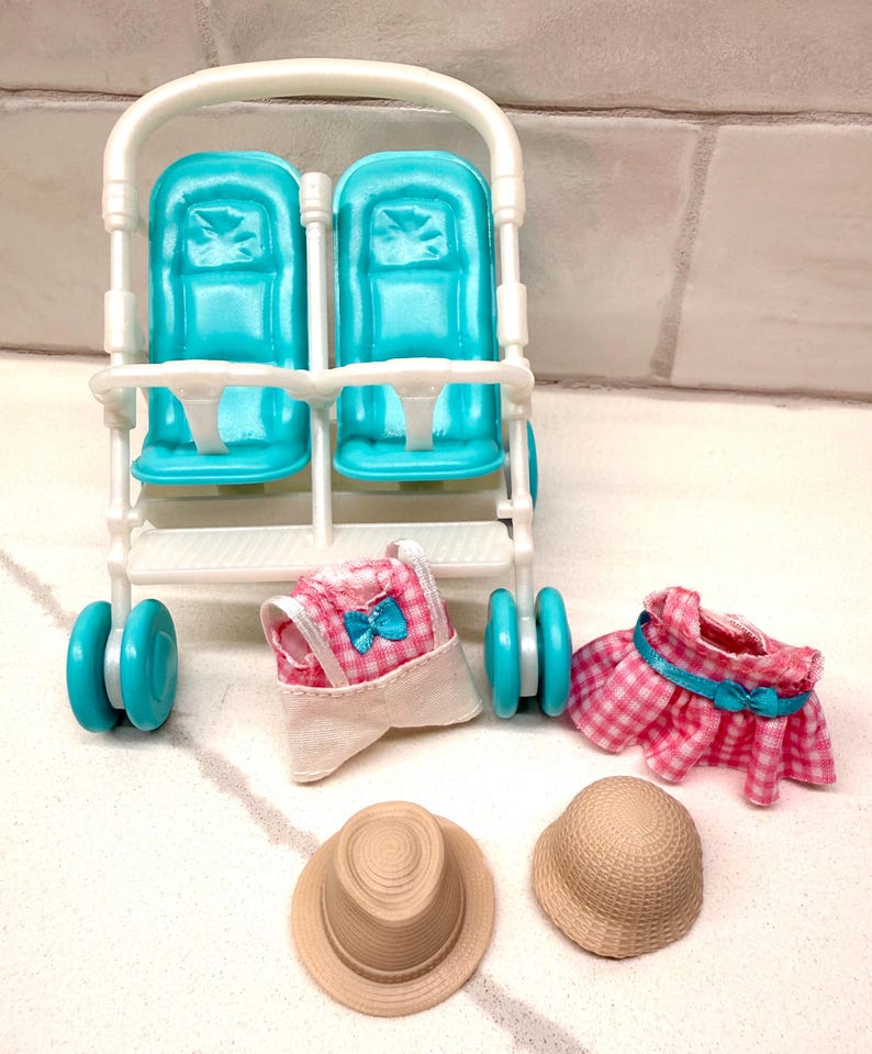 May include: A white and teal double doll stroller with matching teal wheels. Accessories include a pink gingham dress, a pink gingham sun hat, and two beige straw hats.