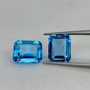 以下が含まれることがあります： エメラルドカットが施された、鮮やかで透明なスカイブルーの宝石2つ。1つは水平に置かれ、もう1つは金属製のピンセットで垂直に保持されており、透明なファセットと明るく反射する表面がはっきりと確認できます。