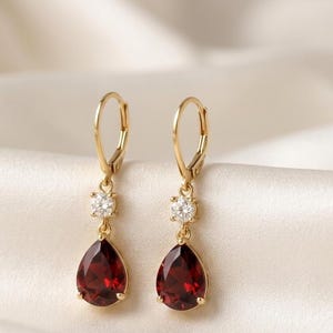 May include: A pair of gold drop earrings featuring a pear-shaped deep red garnet gemstone suspended below a single round brilliant-cut clear stone. The earrings are attached to gold lever-back closures, showcasing a classic design with metal.