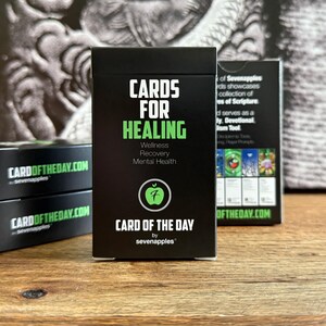 May include: A black deck of cards titled "CARDS FOR HEALING" with green and white text. The deck includes the text "Wellness, Recovery, Mental Health" and "CARD OF THE DAY". Two additional decks are visible, along with a card showcasing images.