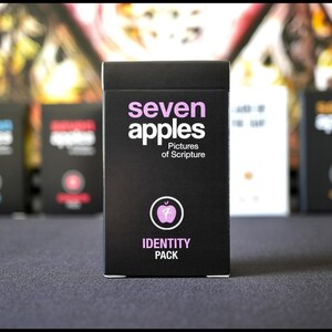 May include: A black box with the text "seven apples" in white and the words "Pictures of Scripture" in smaller white text. The box also has a pink apple with a white "7" inside and the words "IDENTITY PACK" in white.